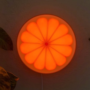 Orange night light glowing bright amber-orange from the front in a dark room, its petal segments lit against a near-black wall. A bold retro citrus mood light and fruit wall art for a bedroom, dorm, or cozy corner.