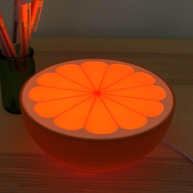 Orange slice lamp glowing warm on a desk in a dark room beside a cup of pens, casting a cozy amber glow. A retro citrus table lamp and mood light for a bedroom, office, or dorm.