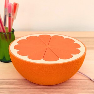 Orange half-dome lamp sitting as a glowing bowl on a wood desk next to a cup of pens. A retro table lamp and citrus night light that works as both wall art and a desk accent for a home office or shelf.