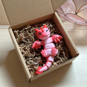 May include: A pink and red articulated dragon figurine with a keychain, nestled in a cardboard box filled with brown paper filler. The dragon has a smiling face and outstretched wings. A unique gift idea.