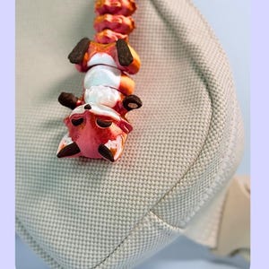 Op de afbeelding: Een Flexi Fox sleutelhanger, een tashanger en een fidget-speeltje. De sleutelhanger heeft een reeks oranje, witte en zwarte vosfiguren. De tekst "Flexi Fox Keychain" is roze. De sleutelhanger is bevestigd aan een zilverkleurige metalen ring.