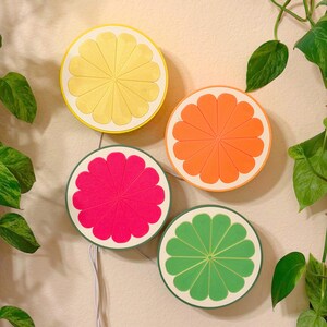 A set of four 3D printed citrus slice LED lamps in lemon, orange, grapefruit, and lime arranged on a pale wall with trailing pothos vines. The fruit-shaped wall lights glow softly in a bright, plant-filled room, working as colorful wall art and gentle ambient lighting. A handmade decor set for a bedroom, kitchen, dorm, or cozy reading nook, and a playful housewarming gift.