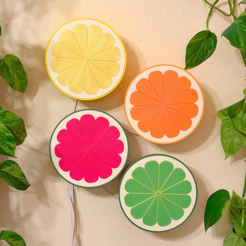 A set of four 3D printed citrus slice LED lamps in lemon, orange, grapefruit, and lime arranged on a pale wall with trailing pothos vines. The fruit-shaped wall lights glow softly in a bright, plant-filled room, working as colorful wall art and gentle ambient lighting. A handmade decor set for a bedroom, kitchen, dorm, or cozy reading nook, and a playful housewarming gift.
