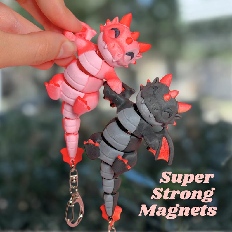 May include: Two articulated dragon magnets, one pink and one gray, are held up by a hand. The dragons have red accents and are attached to keychains. The text "Super Strong Magnets" is visible.