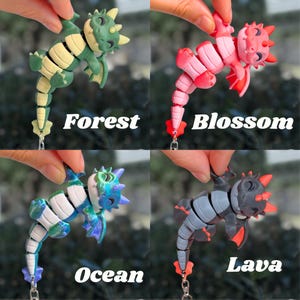 May include: Four articulated dragon figurines in different colors: green and yellow "Forest", pink "Blossom", blue and white "Ocean", and gray and red "Lava". Each dragon is held by a hand and has a keychain attachment. The names of the dragons are written below them.