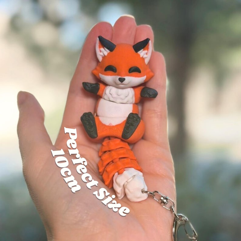 Classic Orange 3D printed articulated fox keychain resting in an open palm showing its perfect 10cm size. The fox sits upright with fluffy curled tail and silver lobster clasp visible. A satisfying size to hold, fidget with, or clip to any bag or backpack. Text reads Perfect Size 10cm. Fox keychain size reference 3D printed cute fox palm size articulated fox fidget magnetic paws