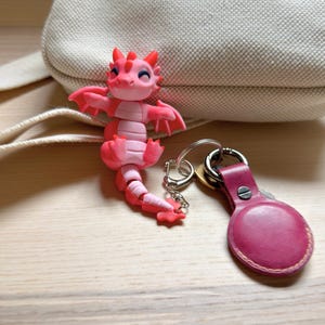 May include: A pink dragon keychain with a matching leather AirTag holder. The dragon is articulated and has a smiling face. The keychain is attached to a silver keyring.
