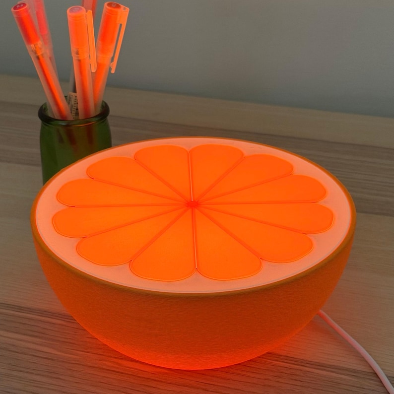 Orange slice lamp glowing warm on a desk in a dark room beside a cup of pens, casting a cozy amber glow. A retro citrus table lamp and mood light for a bedroom, office, or dorm.