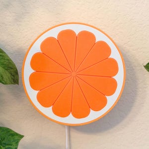 Tangerine orange slice wall lamp lit on a pale wall beside trailing greenery. A cheerful fruit wall art piece and warm night light that brings retro color to a home office, shelf, or colorful room.