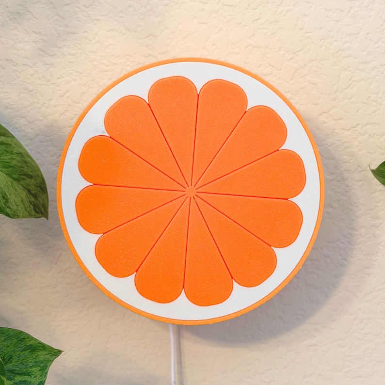Tangerine orange slice wall lamp lit on a pale wall beside trailing greenery. A cheerful fruit wall art piece and warm night light that brings retro color to a home office, shelf, or colorful room.