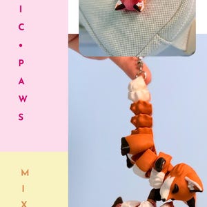 Op de afbeelding: Een fidget-speeltje in de vorm van een vos met een sleutelhanger. De vos is oranje, wit en bruin, met een gesegmenteerd lichaam. De tekst "CUTE &bull; FIDGET" en "MAGNETIC &bull; PAWS" is ook zichtbaar.