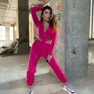 May include: A woman wearing a pink two-piece outfit with a zipper detail. The outfit includes a cropped top and matching pants. She is standing in a concrete building with a sign that reads "Fire Exit".