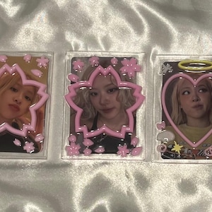 Puede incluir: Tres portatarjetas fotográficas de acrílico transparente con imágenes de una mujer. Cada tarjeta está enmarcada con un borde rosa en forma de flor o corazón. El fondo es una tela blanca y sedosa.