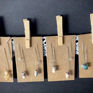 May include: Five pairs of dangle earrings with different colored stones and patterns. The earrings are displayed on brown card stock with wooden clothespins.