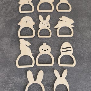 Peut inclure: Une collection de onze ronds de serviette en bois sur le thème de Pâques. Les motifs comprennent des lapins, des poussins et un œuf, tous avec une ouverture circulaire pour une serviette. Les ronds de serviette sont en bois clair.