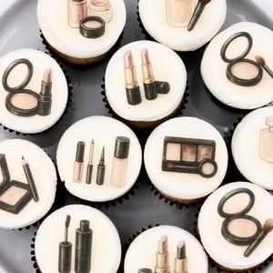 May include: A dozen cupcakes arranged on a gray plate, each topped with a white frosting disc. The discs feature various makeup illustrations, including lipstick, eyeshadow, foundation, and brushes. The cupcakes are a light brown color.