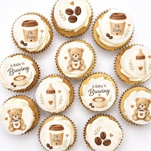 Könnte beinhalten: Eine Ansammlung von elf Cupcakes mit weißem Zuckerguss und runden essbaren Auflegern, die Illustrationen von Teddybären, Kaffeetassen mit dem Text Oh Baby oder A Baby Is Brewing, Babyflaschen und Kaffeebohnen auf weißem Grund zeigen.