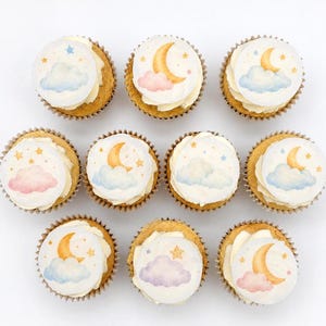 May include: An overhead shot of a dozen cupcakes with white frosting and edible toppers. The toppers feature watercolor designs of a crescent moon, clouds, and stars in pastel colors. The cupcakes are in brown paper liners, arranged on a white surface.