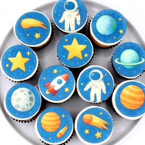 May include: A dozen cupcakes with blue frosting and space-themed decorations. Each cupcake features a different design, including an astronaut, planets, stars, a rocket ship, and a comet. The cupcakes are arranged on a gray plate.