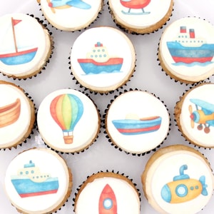 May include: A dozen cupcakes with white frosting, each decorated with a different colorful transportation design. Designs include a plane, helicopter, boat, ship, rowboat, hot air balloon, and submarine. The cupcakes are arranged on a white plate.