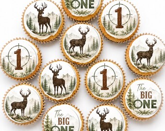 May include: A collection of twelve cupcakes featuring edible toppers with printed designs. The designs include a brown deer in a forest, the number 1 inside a crosshair, and the text The BIG ONE with a crosshair, all set against mountains.