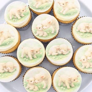 May include: A dozen cupcakes on a gray plate. Each cupcake has a white frosting disc with a lamb illustration on a green grassy background. The lambs are depicted in various poses, some standing and others cuddling.