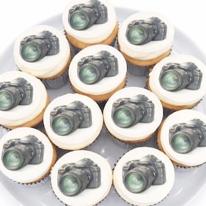 May include: A dozen cupcakes arranged on a gray plate. Each cupcake has a white frosting top with a black and green camera image. The cupcakes are vanilla flavored and are perfect for a photographer.