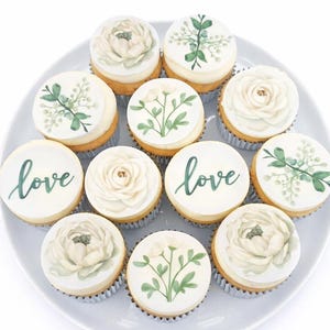 May include: A dozen cupcakes on a white plate. Each cupcake has a white frosting design of flowers or greenery. Some cupcakes have the word "love" written in green script. The cupcakes are in silver cupcake liners.
