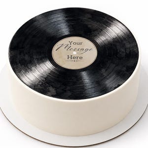 May include: A round cake with smooth white sides and a top decorated to resemble a black vinyl record. The centre label is beige with the text Your Message Here printed in a dark script font, featuring a small decorative line element below.