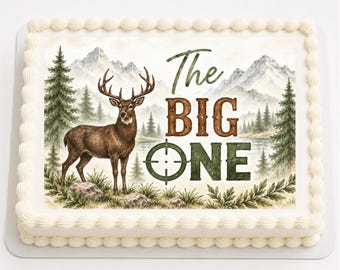May include: A rectangular sheet cake with white piped frosting borders. The center features an edible print of a brown deer standing in a forest with mountains in the background. The text reads The BIG ONE with a green crosshair target symbol.