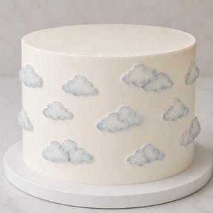 Può includere: Una torta cilindrica con glassa bianca liscia decorata con molteplici ritagli piatti di nuvole color azzurro in stile acquerello applicati all'esterno. La torta poggia su una base circolare bianca, presentando un aspetto pulito e semplice.