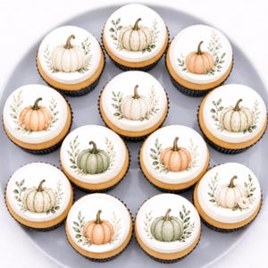 May include: A collection of eleven cupcakes arranged on a gray plate, each topped with a white circular edible topper featuring a watercolor illustration of a single pumpkin in shades of orange, green, or white, surrounded by delicate foliage.