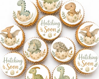 May include: A collection of eleven cupcakes topped with circular edible toppers featuring watercolour illustrations of various baby dinosaurs hatching from spotted eggs and two toppers with the text Hatching Soon in brown script.