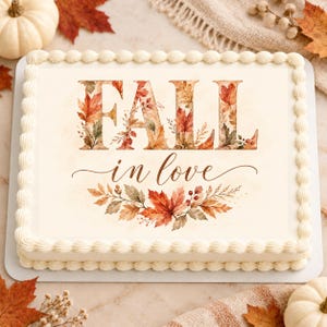 May include: A rectangular sheet cake with white frosting borders featuring the text FALL in love. The word FALL is filled with a watercolor pattern of orange, red, and brown autumn leaves. Below, the words in love are written in brown script.