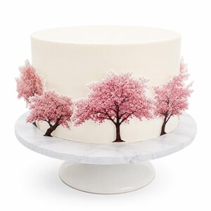 May include: A round cake with smooth white frosting decorated with several flat, printed cherry blossom tree cutouts in shades of pink and brown. The cake sits on a white pedestal stand with a subtle grey marble pattern on the top surface.
