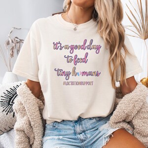 May include: Cream-colored t-shirt with the text "it's a good day to feed tiny humans" in purple and blue script. The shirt also includes the hashtag #LACTATIONSUPPORT. The shirt is a casual style.