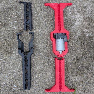 May include: Two disassembled mechanical components. One is black and has a series of decorative cutouts. The other is red and contains a small motor. Both appear to be 3D-printed and are lying on a concrete surface.