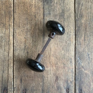 May include: Black ceramic door knob with a metal stem. The knob is on a wooden surface.