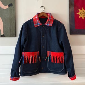 May include: A dark blue denim jacket with a red and blue plaid collar and cuffs. The jacket features two front pockets with red fringe detailing. The jacket is hanging on a wooden hanger.