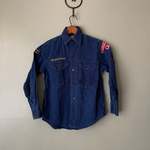 May include: A blue long-sleeved shirt with a button-up front and two pockets. The shirt has a patch that says "CUB SCOUTS B.S.A." and other patches on the sleeves.