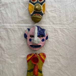 May include: Three colourful, handmade paper masks. The top mask is dark blue with yellow and green accents. The middle mask is pink and blue. The bottom mask is yellow, red, and green. All masks have unique shapes and designs.