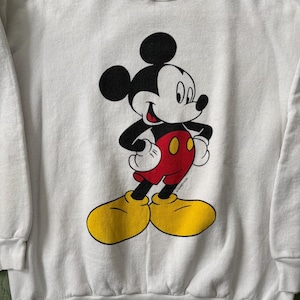 May include: White sweatshirt featuring a classic Mickey Mouse graphic. The cartoon character is depicted in black, red, yellow, and white. Mickey Mouse is standing with his hands on his hips. The sweatshirt has a crew neck and long sleeves.