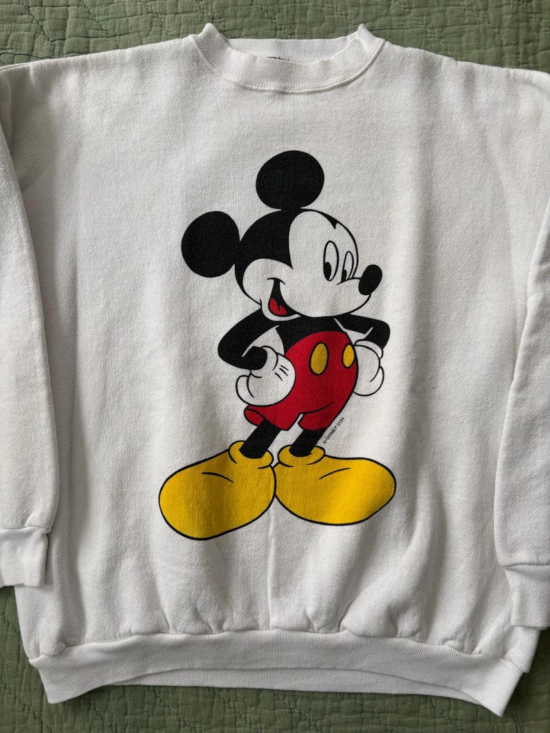 May include: White sweatshirt featuring a classic Mickey Mouse graphic. The cartoon character is depicted in black, red, yellow, and white. Mickey Mouse is standing with his hands on his hips. The sweatshirt has a crew neck and long sleeves.