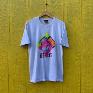 May include: White t-shirt with a colorful graphic of Hershey's Kisses and Reese's Peanut Butter Cups. The graphic is in a diamond shape with the Hershey's logo at the bottom.