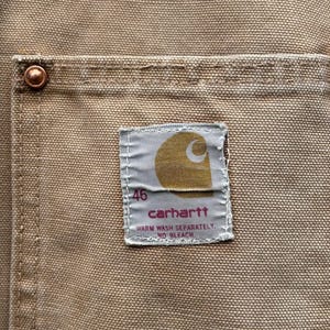 Peut inclure: Gros plan sur une poche en tissu beige avec un rivet en cuivre. Une &eacute;tiquette rectangulaire affiche le logo Carhartt, le chiffre "46" et les instructions de lavage. Le tissu a un aspect textur&eacute;.