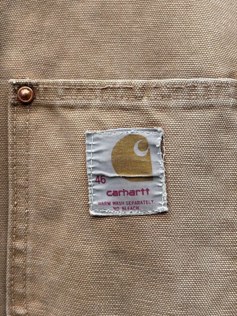 Peut inclure: Gros plan sur une poche en tissu beige avec un rivet en cuivre. Une &eacute;tiquette rectangulaire affiche le logo Carhartt, le chiffre "46" et les instructions de lavage. Le tissu a un aspect textur&eacute;.
