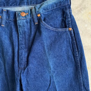 Puede incluir: Un par de pantalones vaqueros de mezclilla azul oscuro con un bot&oacute;n y remaches de cobre. Los vaqueros tienen un estilo cl&aacute;sico de cinco bolsillos.