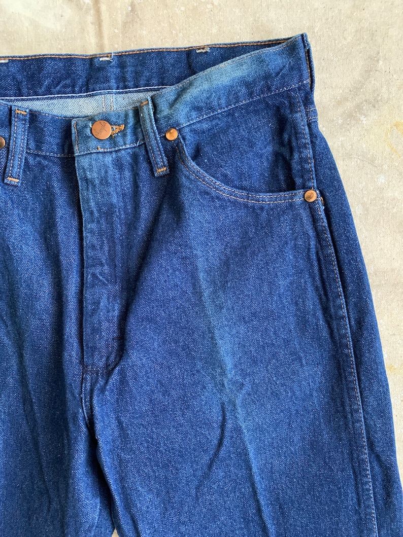 Puede incluir: Un par de pantalones vaqueros de mezclilla azul oscuro con un bot&oacute;n y remaches de cobre. Los vaqueros tienen un estilo cl&aacute;sico de cinco bolsillos.