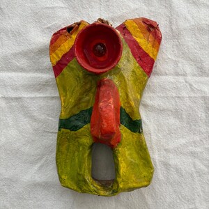 May include: A vibrant, handmade paper mache mask in red, yellow, and green. The mask has a circular red feature at the top, a red nose, and a rectangular opening at the bottom. The mask is displayed against a white backdrop.