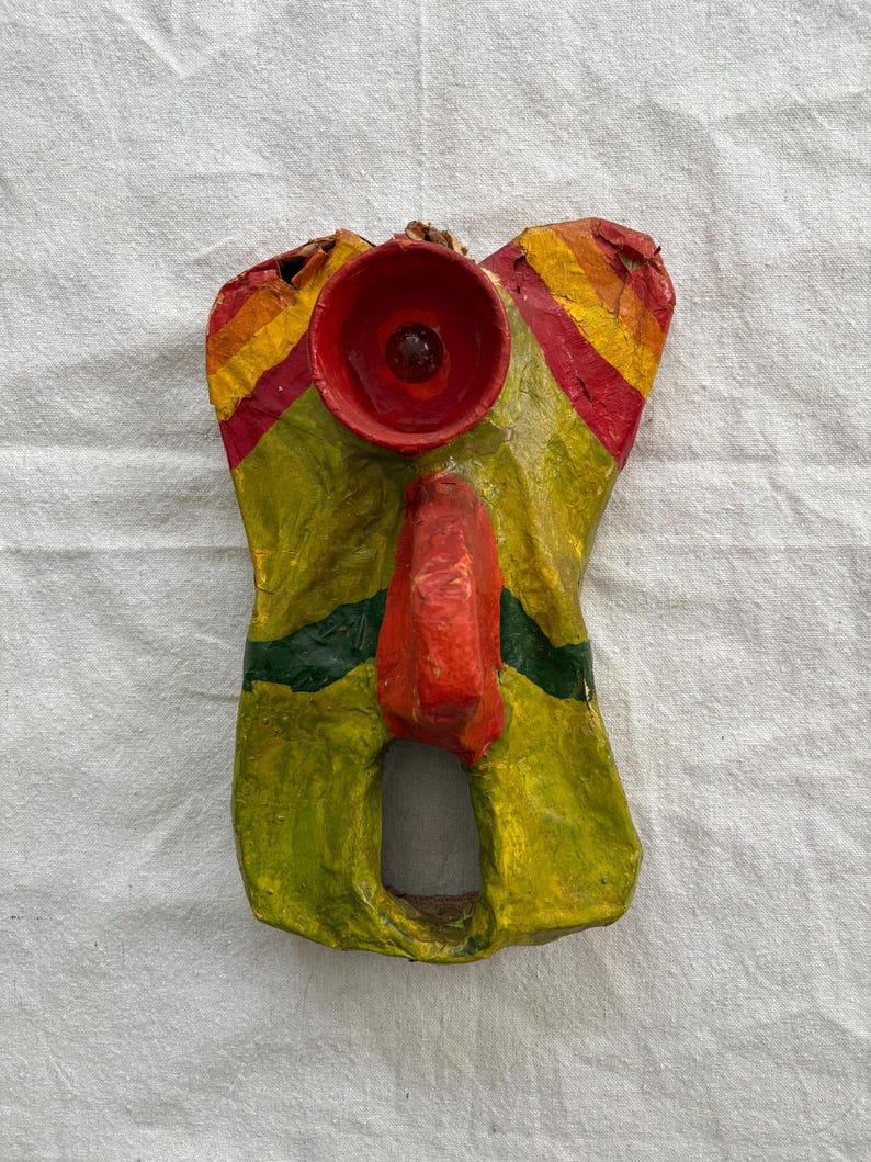 May include: A vibrant, handmade paper mache mask in red, yellow, and green. The mask has a circular red feature at the top, a red nose, and a rectangular opening at the bottom. The mask is displayed against a white backdrop.